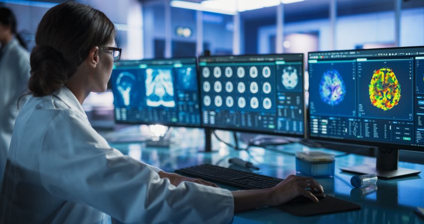 Medical Research Center Laboratory: Female Neurologist Using Desktop Computer With MRI And PET Scans Of a Brain.