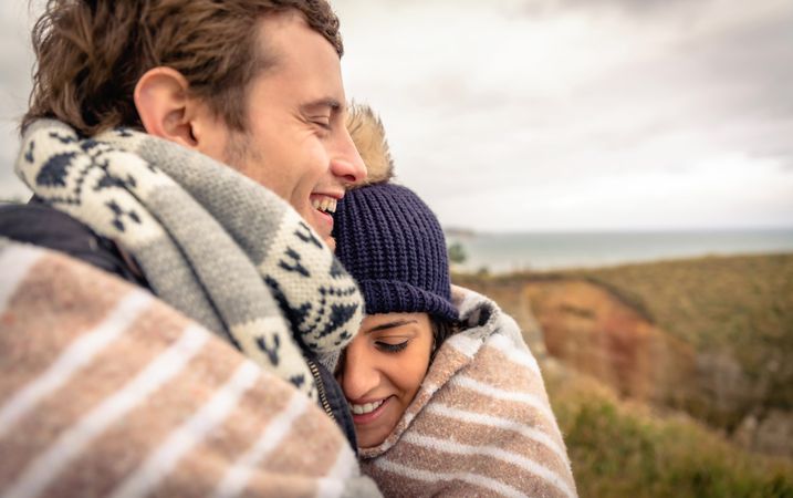 Side view of smiling couple wrapped in blanket on a windy day on a cliff above the coast