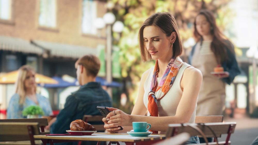 Portrait of a Happy Female Using Smartphone on a Restaurant Terrace, Enjoying a Cup of Coffee. Young Woman Smiling When Receiving a Text, Chatting with Friends Online, Checking Social Network Updates