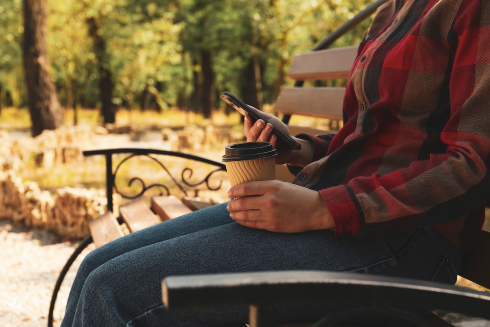 Woman in jeans and shirt hold paper cup and phone outdoor