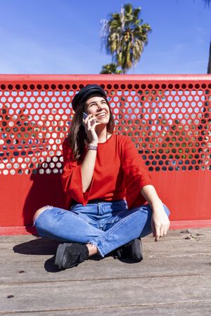 Happy woman in red sitting cross legged talking on mobile phone outdoors on a bright day