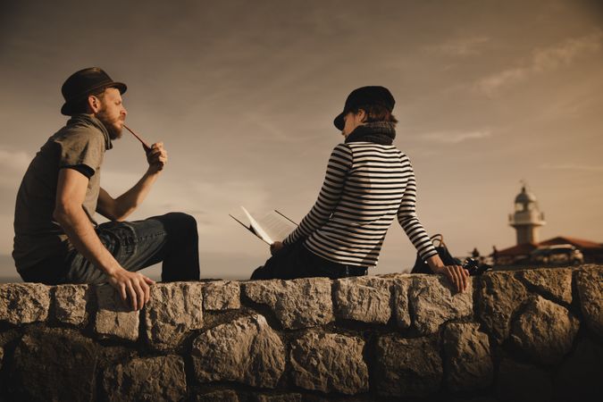 Man with pipe near woman with book on stones near lighthouse