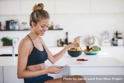 Smiling young woman sitting in kitchen using mobile phone and eating fruits bDV3y5