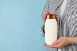 Woman holds blank shampoo bottle on blue background, space for text