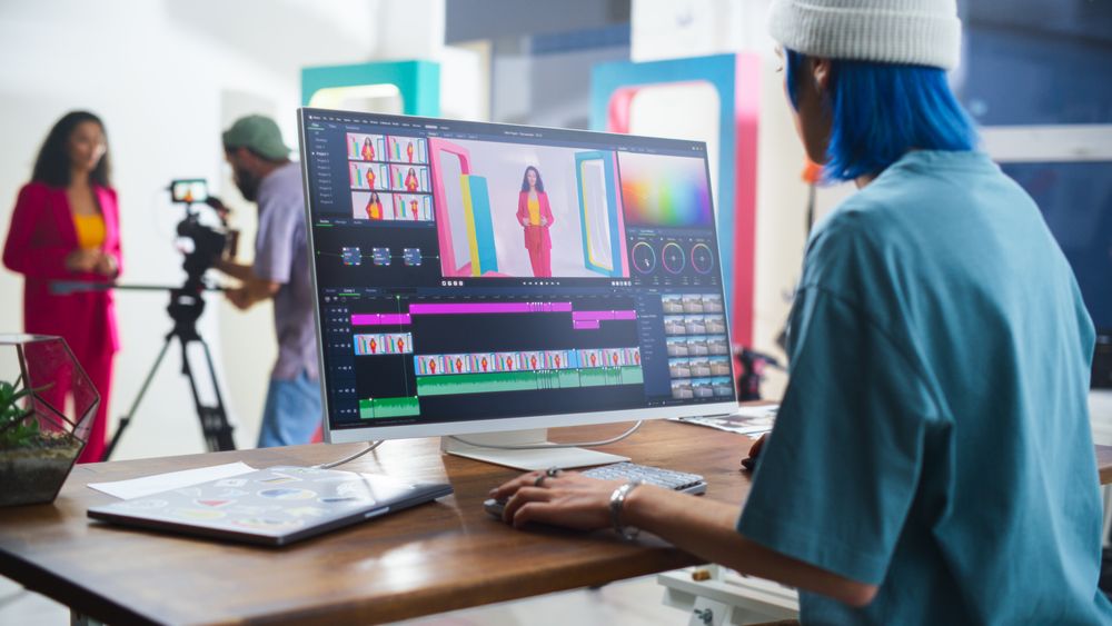 Young White Female Editor With Blue Hair Works On Video Editing Software, While A Hispanic Male Videographer Records A Black Female Host In A Vibrant Studio Setting, Enhancing Media Production.