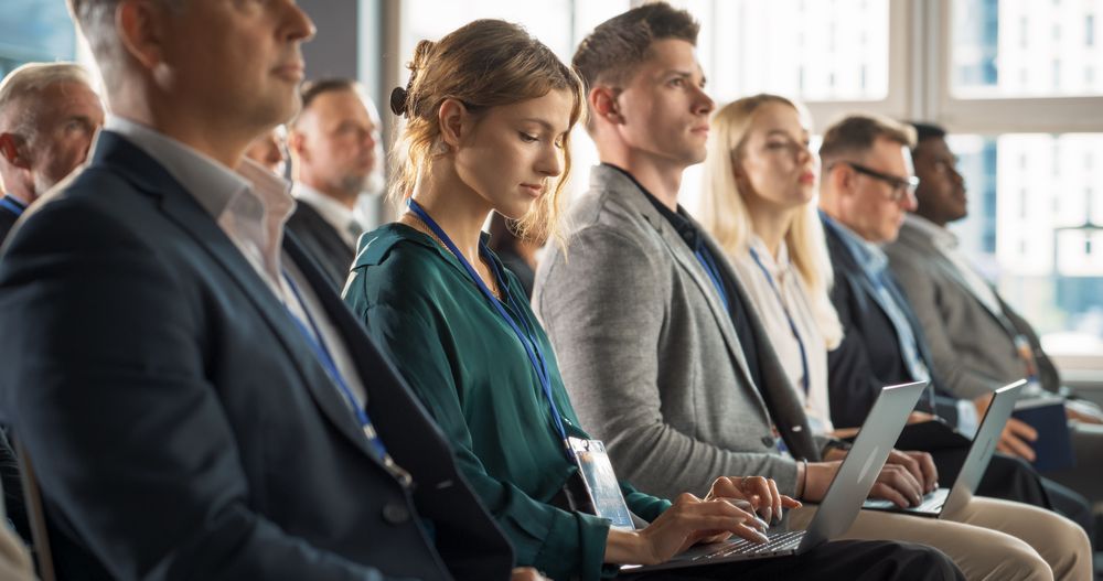Young Woman Sitting in Crowded Audience at Business Conference. Female Delegate Smiling, Using Laptop Computer. Manager Watching Motivational Presentation About Investing In Innovative Startups.