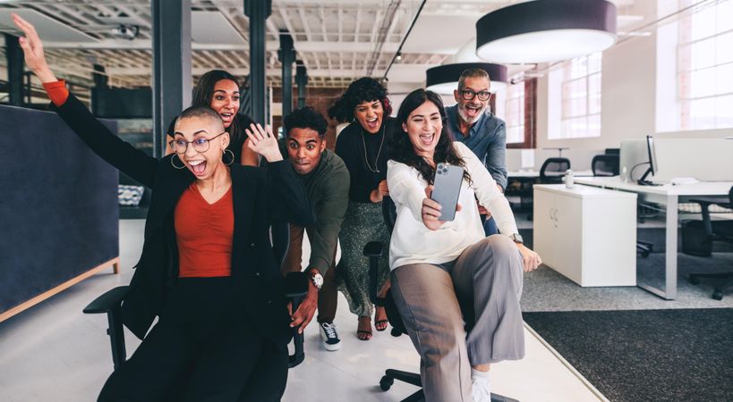 Cheerful businesswoman taking a selfie with her coworkers  while having office chair race at break