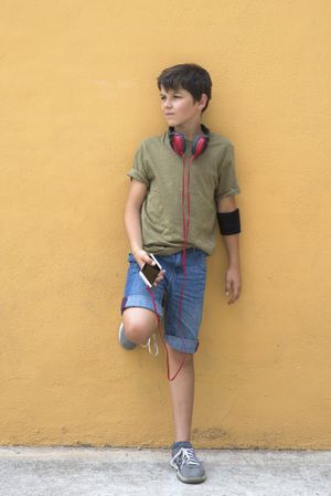 Young boy leaning on a yellow wall with headphones on neck, holding a smartphone