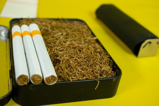 Close up of cigarettes resting on box of loose leaf tobacco on yellow background