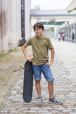 Young boy standing on cobblestone street while leaning on skateboard