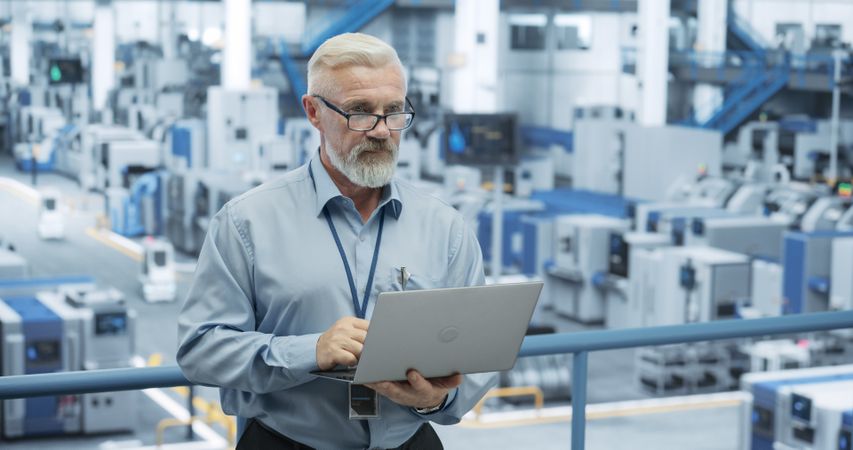 Mature Factory Manager Working on Laptop at a Futuristic Industrial Manufacture with Autonomous AI Robots. Specialist Monitoring All the Data on a Computer, Checking Reports and Planning Workload