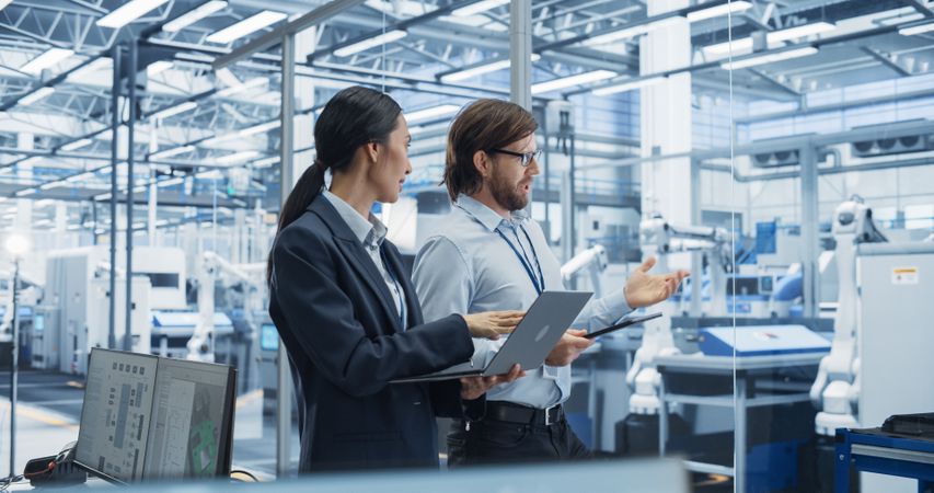 Male Engineer and Female Project Manager Standing in Office, Talking, Using Computers and Monitoring Production at a Modern Automated Electronics Manufacture with AI Robotic Arms