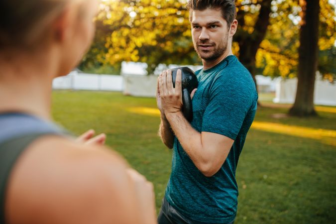 Shot of fit young man exercising with kettlebell outdoors in the park and looking at woman