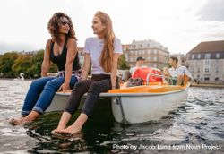 Shot of two women sitting in front pedal boat with feet in water and man in background 4BV2x4