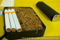 Close up of cigarettes resting on box of loose leaf tobacco on yellow background 4AzmDQ