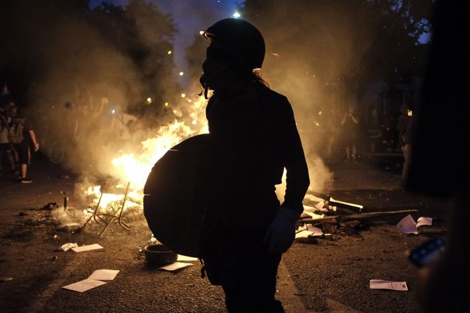Person with helmet and wooden shield beside fire at protest