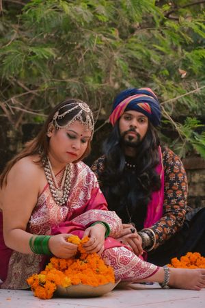 Man with long hair wearing a turban sitting beside woman in pink sari on ground beside trees
