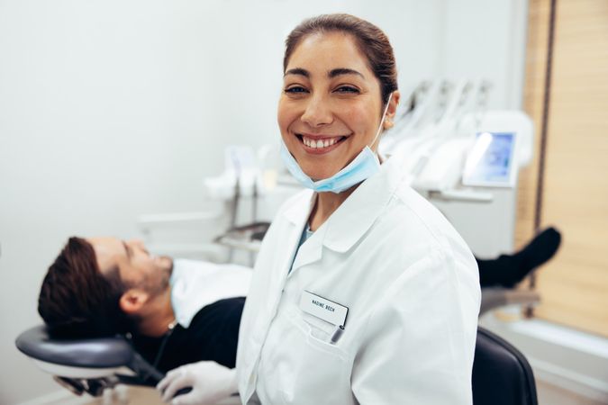 Happy dentist smiling at camera with male patient in the background