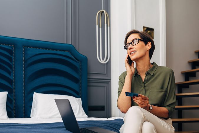 Woman sitting on hotel room bed with phone, laptop and credit card