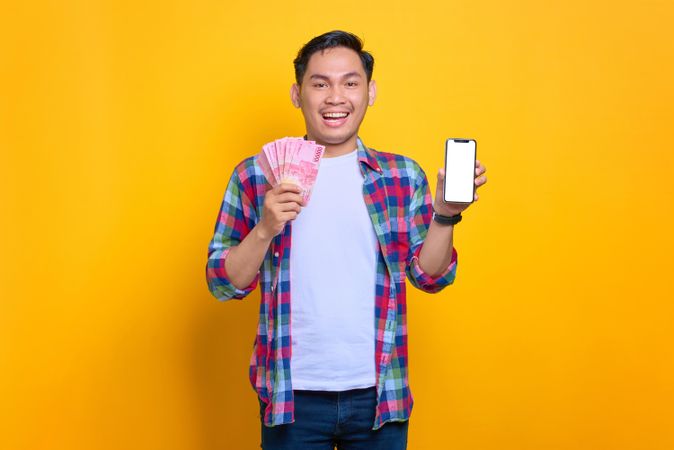 Smiling Asian man holding cash and showing blank phone screen in studio shoot