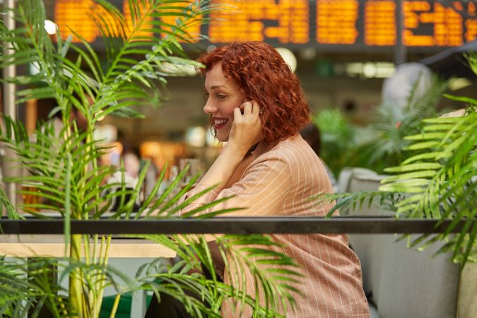 Cheerful young woman working on laptop in cafe and talking phone