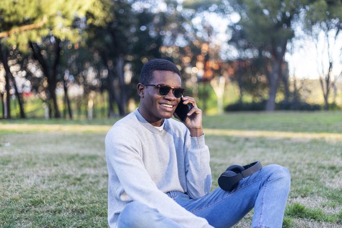 Happy Black male with sunglasses sitting in park talking on his phone