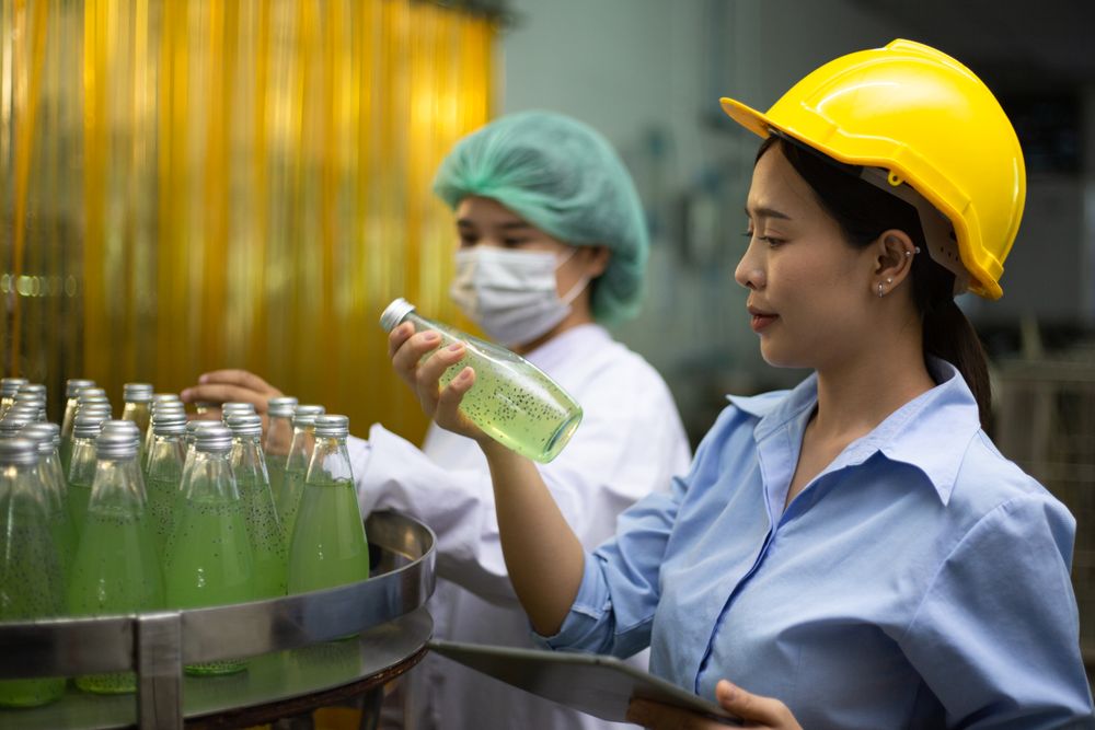 Asian female manager and worker holgin digital tablet checking information and quality control for juice drink product on bottling factory machinery.