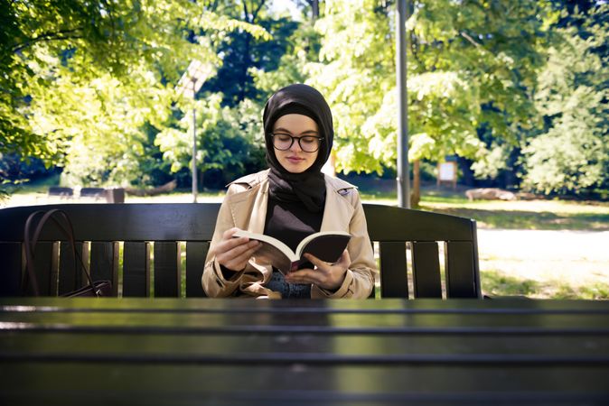 Woman in headscarf sitting at picnic table in park with a book