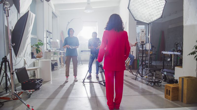 Young Diverse Team Of Filmmakers In A Bright Studio Setting, With A Woman In A Red Suit Directing The Shoot, Surrounded By Cameras, Lights, And Crew Members Focused On Creating Engaging Content.