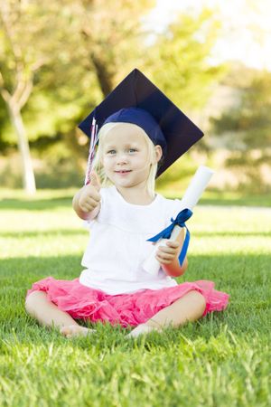 Little Girl In Grass Wearing Graduation Cap Holding Diploma With Ribbon