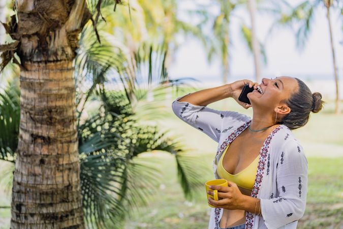 Cheerful woman talking on phone while drinking coffee by palm tree
