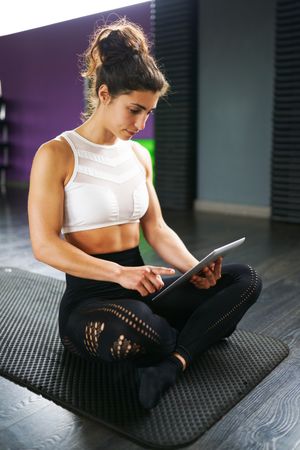 Fit woman cross legged on yoga mat looking down at a digital tablet, vertical