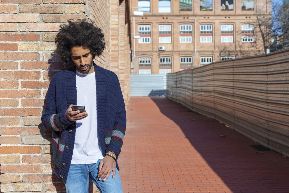 Handsome Black man using his smartphone with smile while leaning on a bricked wall outdoors on sunny day