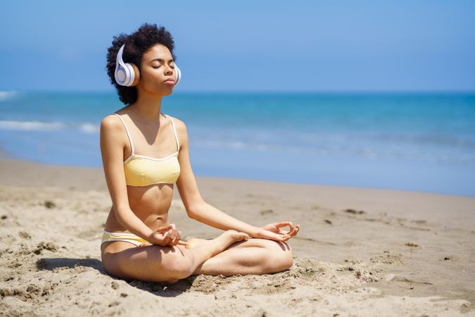 Woman in yellow swimwear sitting cross legged and meditating on sandy beach in head phones