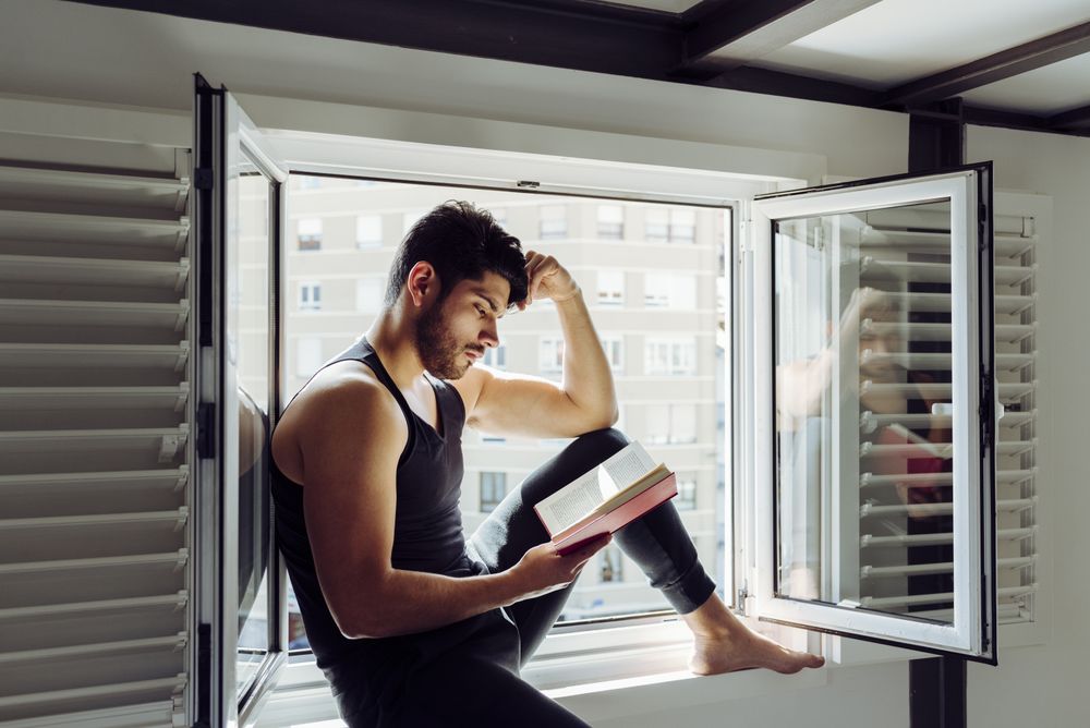 Young male sitting on window and reading book