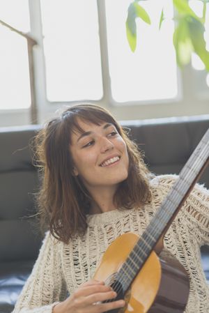 Happy female in wooly sweater looking at fretboard of guitar