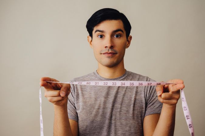 Serious Hispanic male holding measuring tape between hands in beige studio shoot