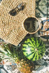 Thatched bag laying on pattered tiles with sunglasses, watermelon and pineapple