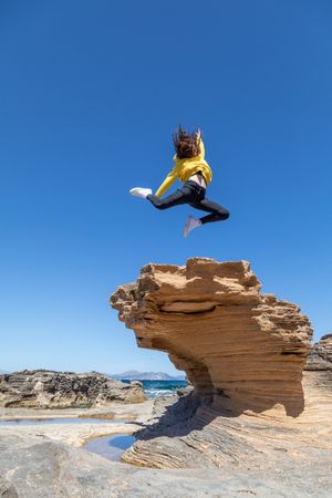 Sportive woman jumping above rock against blue sky
