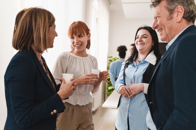 Group of colleagues in smart casual attire conversing and laughing near a window during an office break