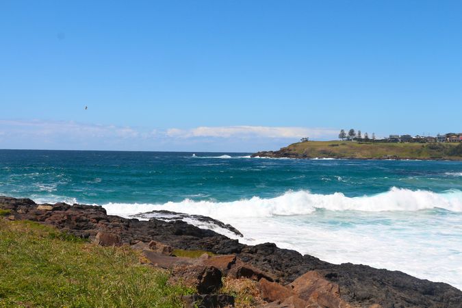 Waves crash against rocky shoreline under a clear blue sky near coastal settlement