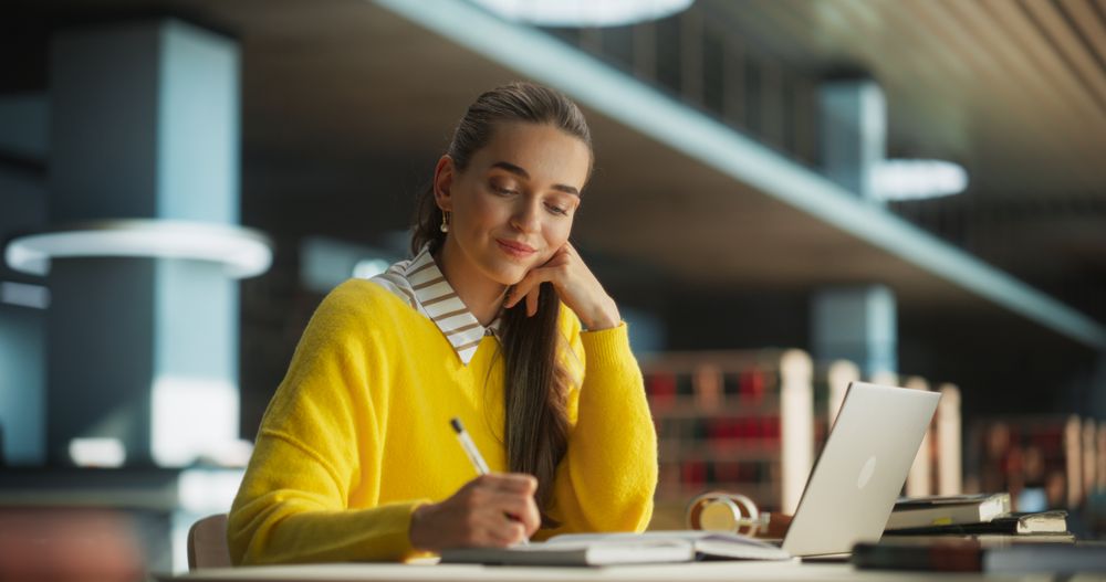 Student Writing a Social Science Essay on a Laptop Computer and Taking Notes in a Paper Notebook. Young Stylish Female Studying Alone in a Public Library
