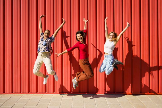 Three women happily jumping in front of red wall on sunny day