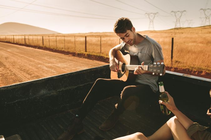 Young male musician playing guitar in cargo bed of truck while girlfriend holds beer bottle