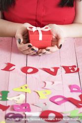 Woman’s hands holding a red gift box over a pink wooden table full of multicolor paper letters 5aMp85