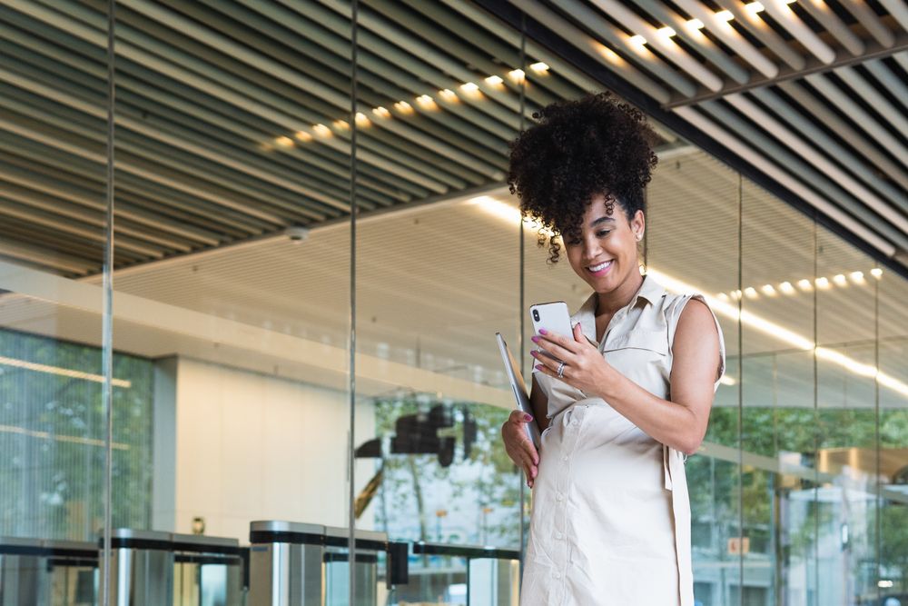 Smiling Black pregnant woman using smartphone and carrying laptop