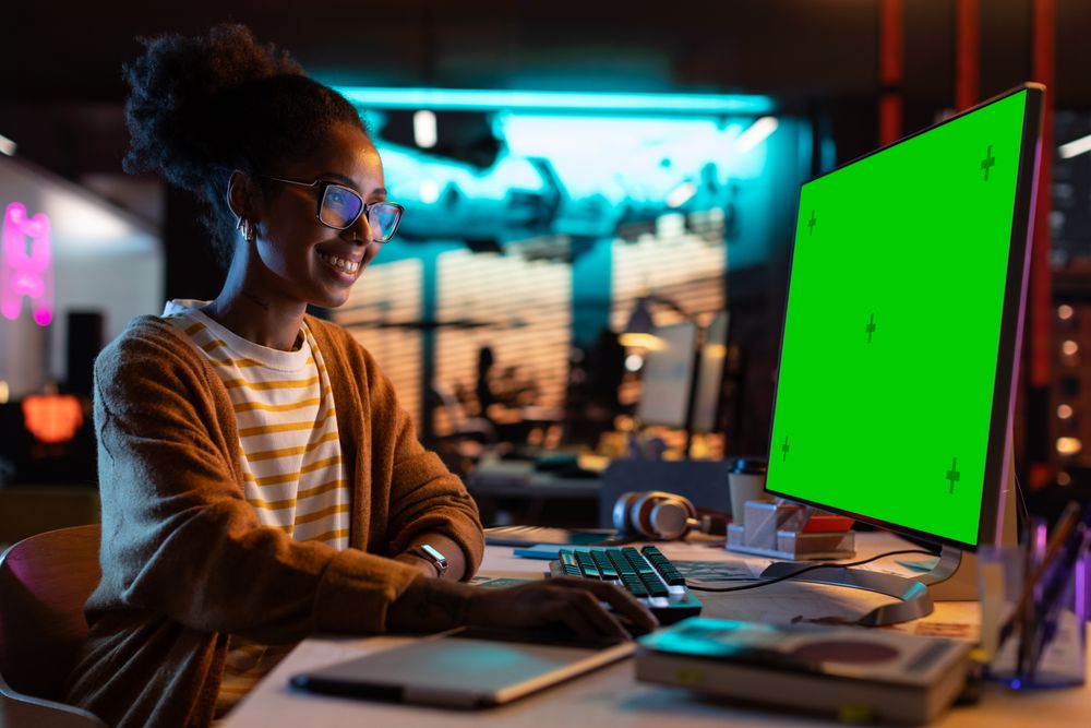Portrait of Creative Black Woman Sitting at Her Desk Using Desktop Computer with Mock-up Green Screen.