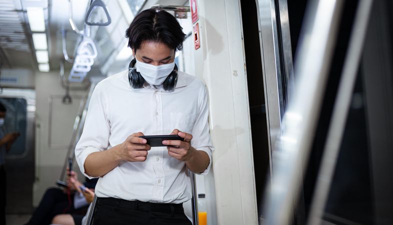 Man in facemask and headphones looking at smartphone in metro car