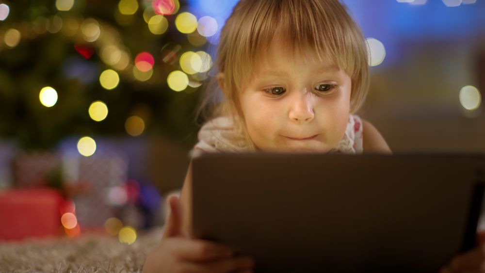 Little Girl With Tablet Computer in Her Hands Lies on the Carpet under the Christmas Tree.