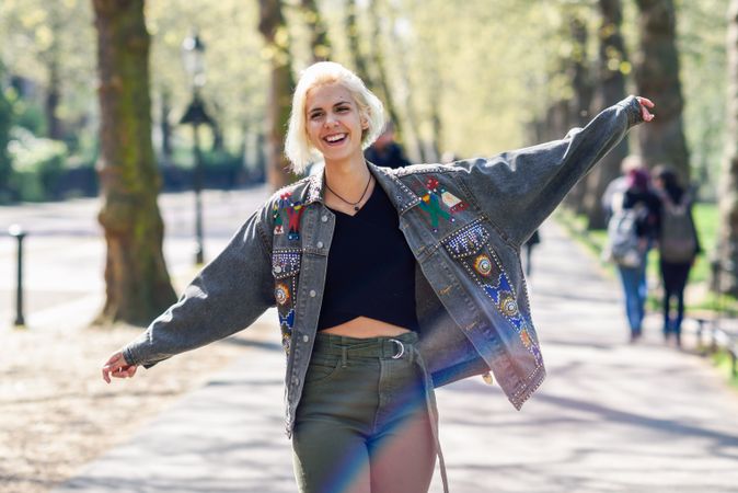 Blonde woman smiling in jean jacket with arms outstretched while walking in park